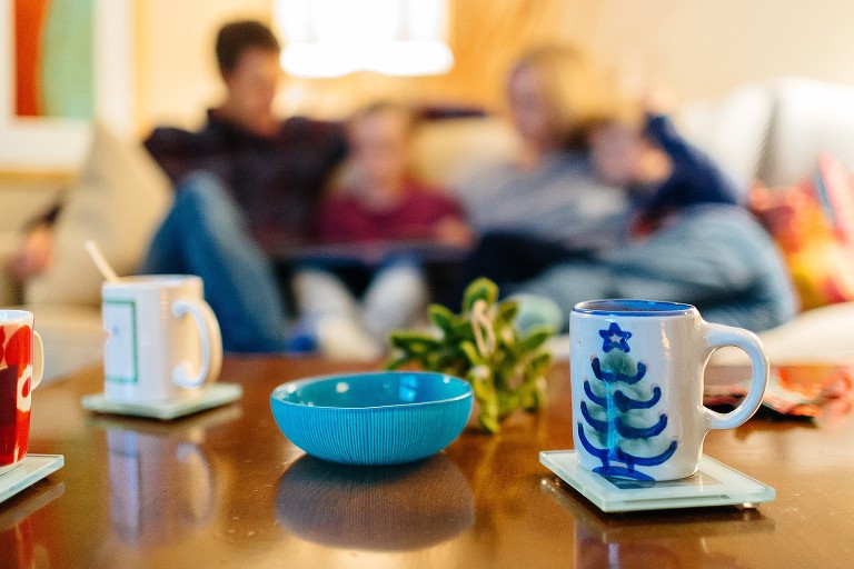 Focus on mugs of hot chocolate while family is in the background reading Christmas story on the couch 