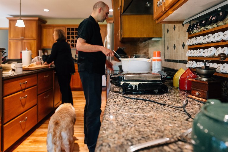 The waffle iron steams and waffle batter drips batter while Dad prepares bacon and Mom washes fruit. The family dog walks through the scene. 