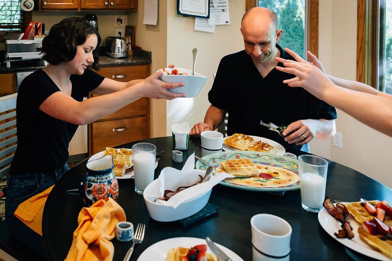 Daughter passes bowl of fruit to sister over a crowded breakfast table. Sister's hands are outstretched. 