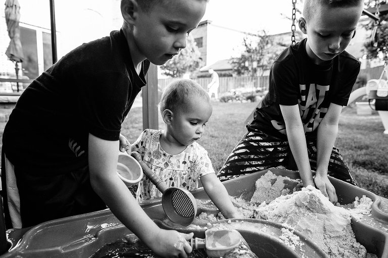 Three siblings play in sand box, black and white 