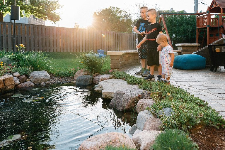 Three siblings throw small stones in backyard pond. 