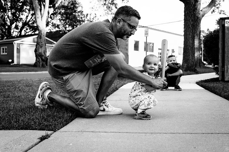 Dad helps toddler girl hold bat, while brother holds catcher mitt behind them