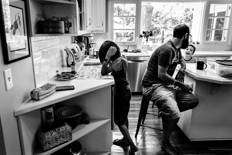 Black and white. Dad laughs with preschool son, older son reads the Sunday comics. 