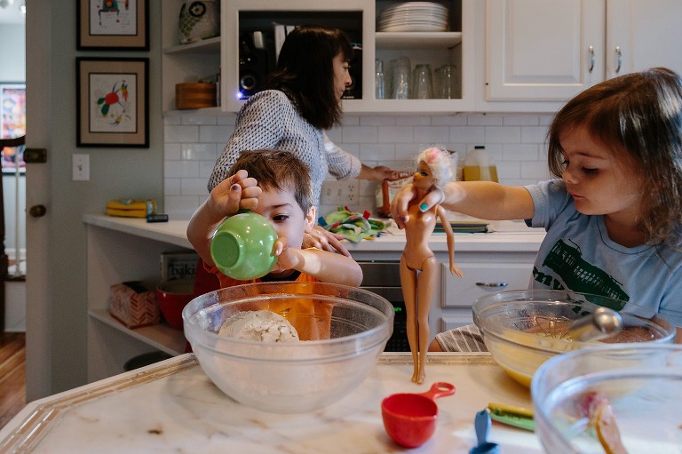 Toddler boy dumps flour in glass bowl while Mom returns flour container to counter and toddler sister watches. 