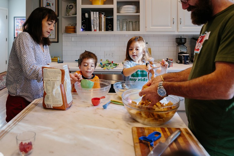 Mom, two toddlers, and Dad work together in the kitchen. Mom and son pour flour in a glass bowl, Dad tosses apples with brown sugar mixture. 
