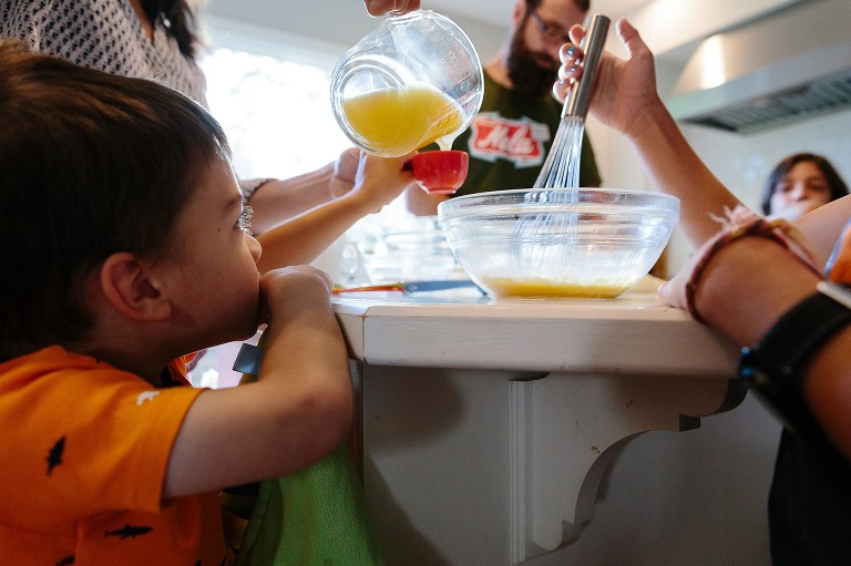 Youngest son holds measuring spoon for orange juice. Older son whisks. 