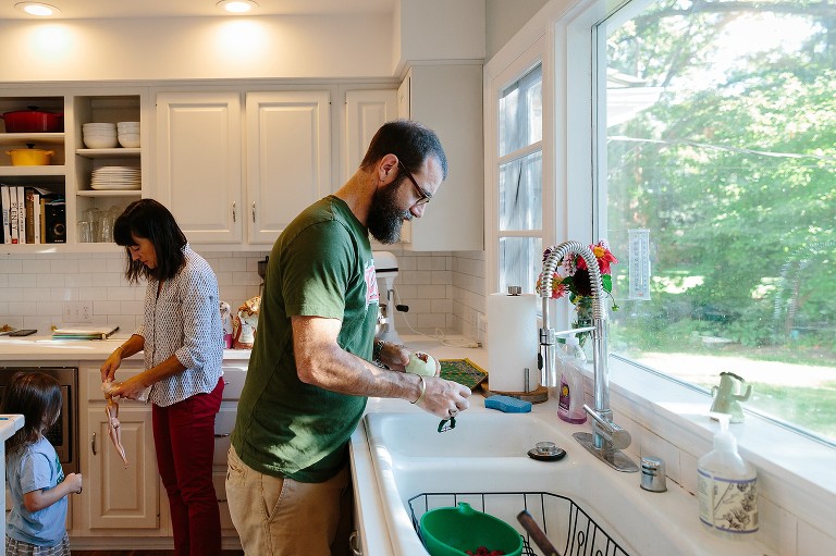 Dad peels apples at sink, Mom ties Barbie's hair for preschooler 
