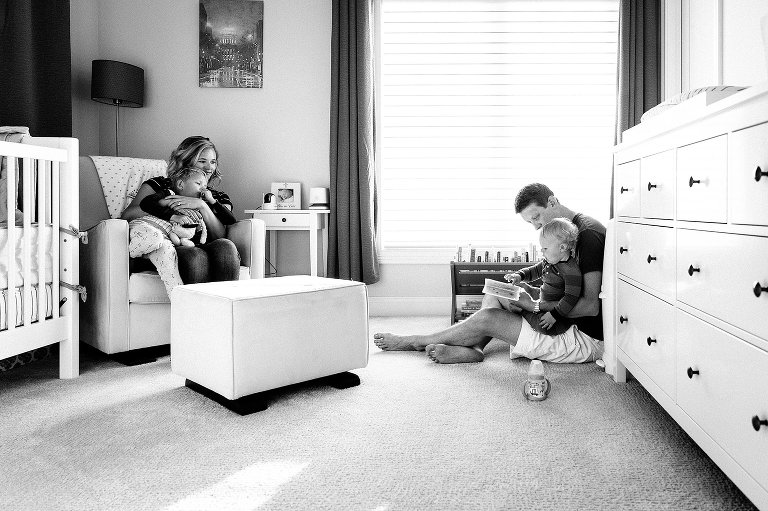 Dad reads book to toddler son while mom and preschool son listen from chair 