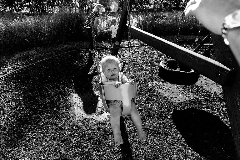 Dad's hand pushes toddler boy in swing while mom and preschool boy are in background.