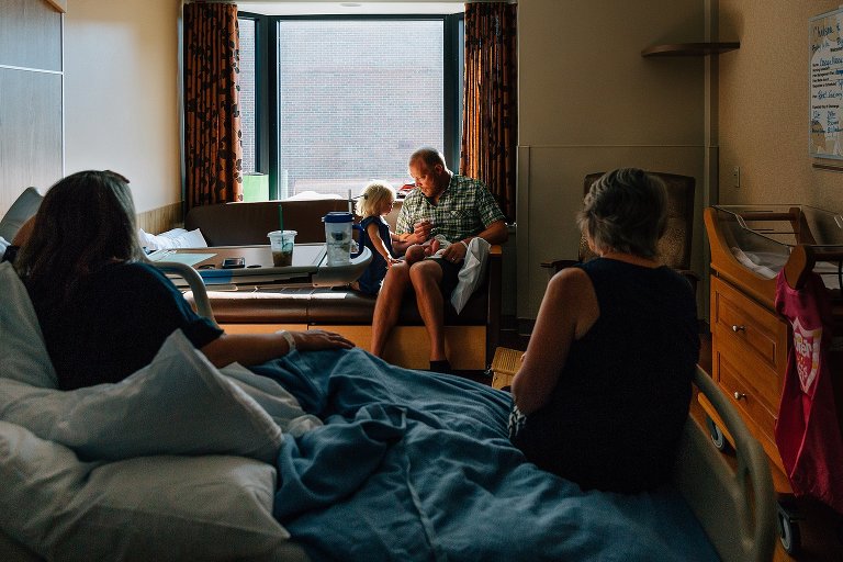 Dad, toddler, and newborn sit in hospital window while mom and grandma look on