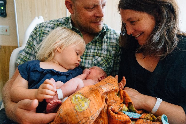 Mom looks at toddle daughter, toddler daughter looks at newborn sister, Dad looks at Mom.