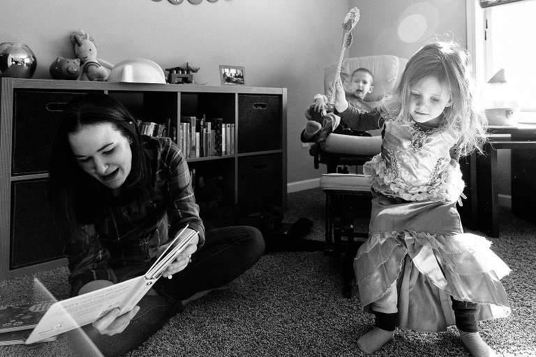 Mom reads to baby off camera, boy lounges in chair, daughter leaps forward wearing dress up clothes 