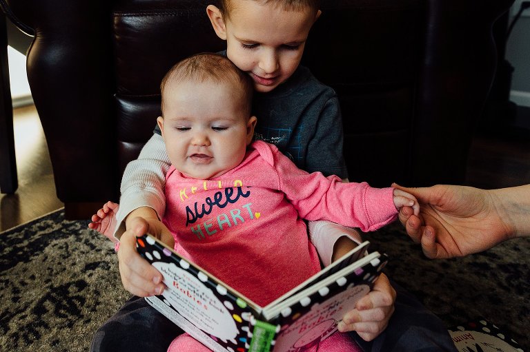 Baby sister sits in brother's lap while he reads to her. 