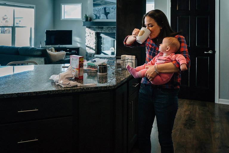 Mom drinks coffee while holding baby; baby looks on. 