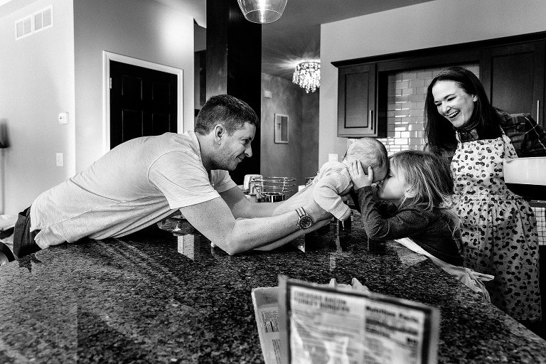 Dad stretches across kitchen island wiht baby so baby and older sister can have a kiss. 
