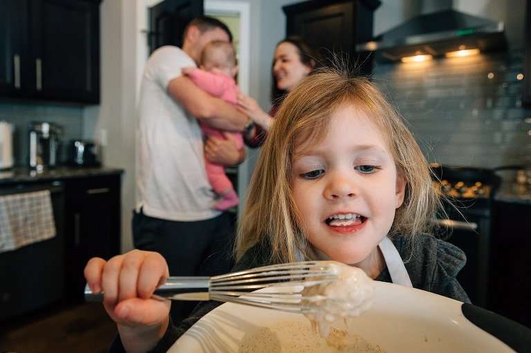 Daughter sneaks a bit of pancake batter while parents are engaged with baby in background 