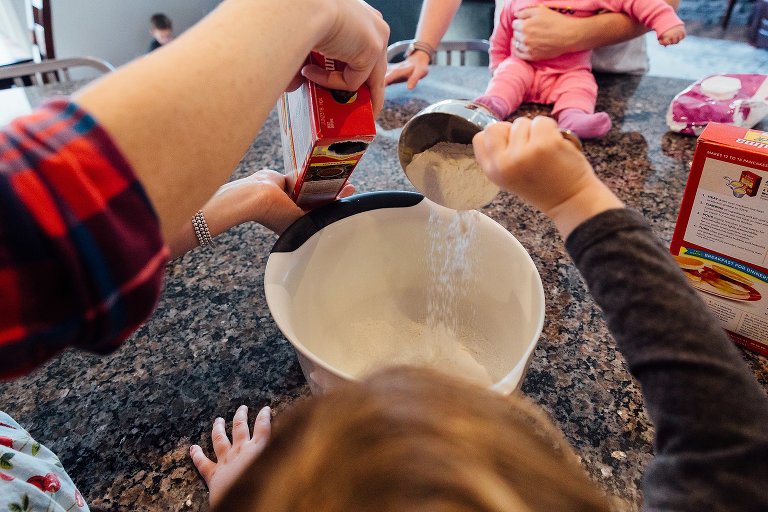 Young girl dumps pancake mix into bowl. Baby watches. 