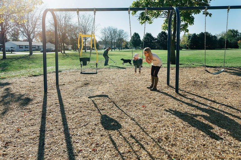 Mom gently pushes baby on swing, while dad plays catch with dog in background