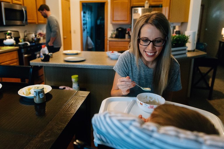 Mom smiles at baby while holding a spoon of baby cereal. Dad is in the background making pancakes