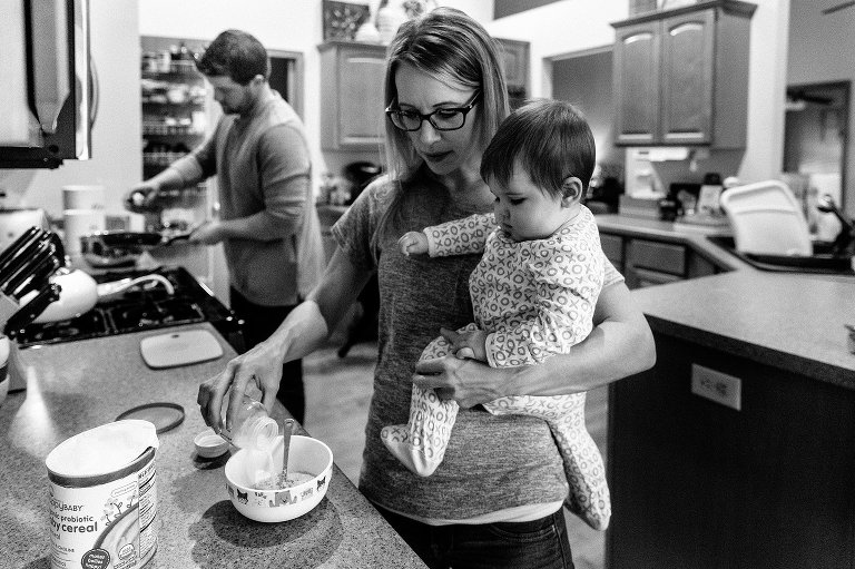 Mom prepares baby cereal, while holding baby in arm. Dad prepares pan of pancakes in background.