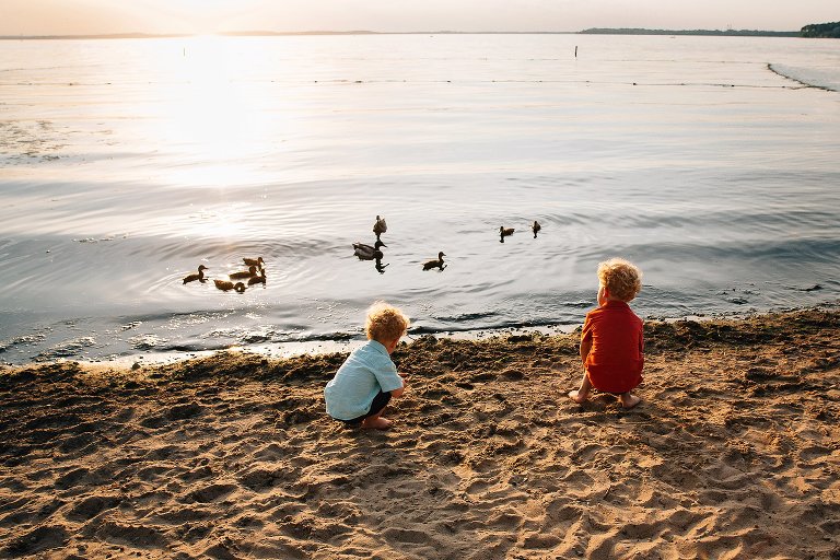 Young boys play in sand and watch ducks in the lake. 