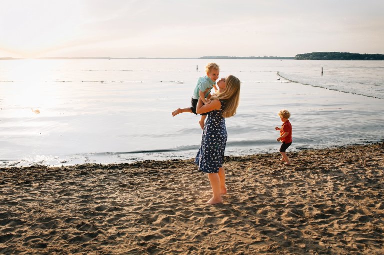 Mom scoops up young son while older son plays in the background. 