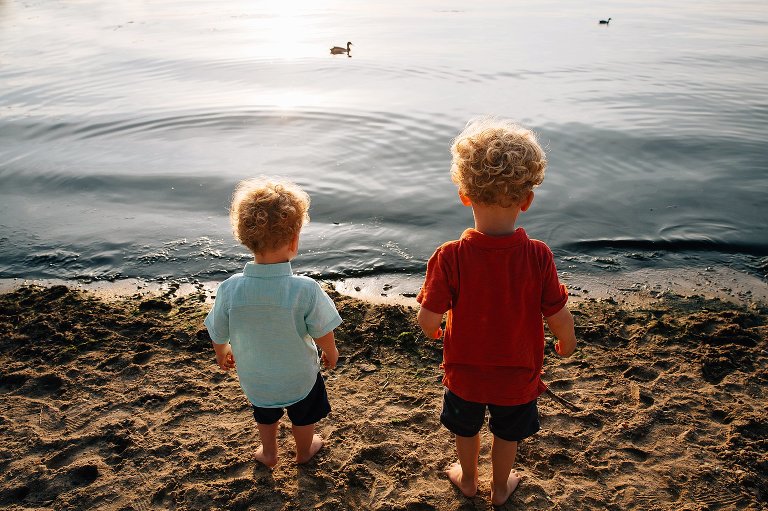 Two young boys stand at water's edge as ducks swim past.