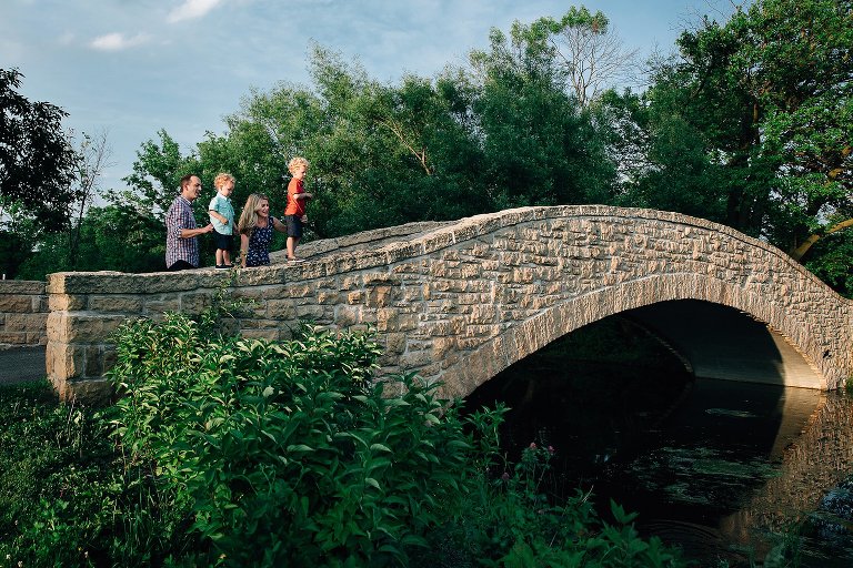 Family stands on bridge and toss rocks in lake. 