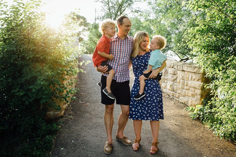 Family stands at the base of a bridge. Mom and Dad hold young sons and are engaged with them. 
