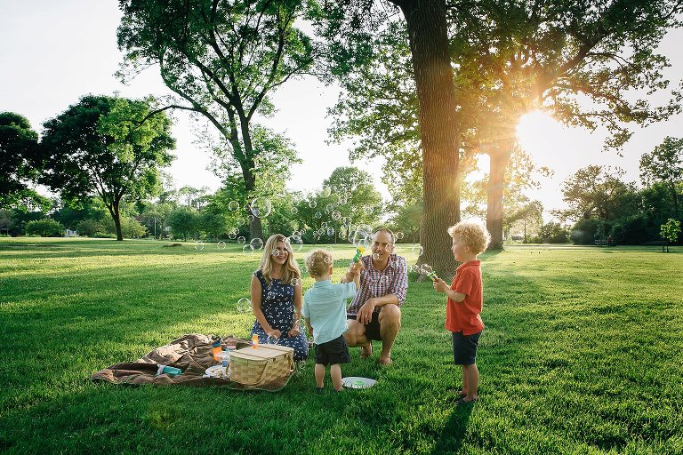 Family of four sits in the park, having a snack and blowing bubbles. Summer sun is low.