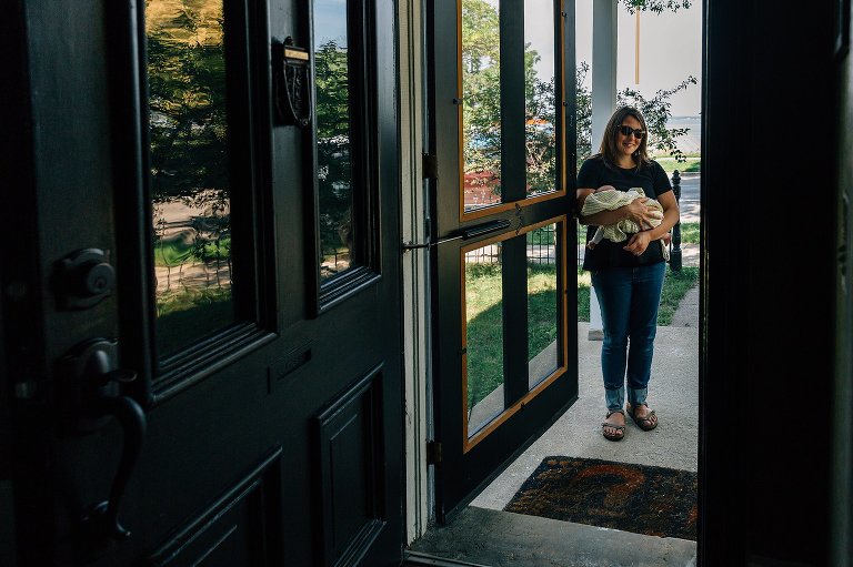 Mom stands in the doorway of house wiht newborn daughter. Lake in the background. 