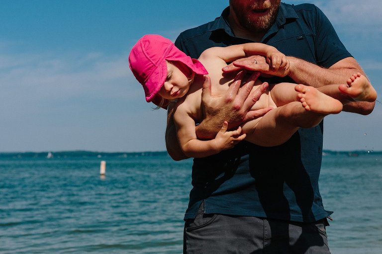 Dad rinses child in lake. High shadows indicate time of day. 