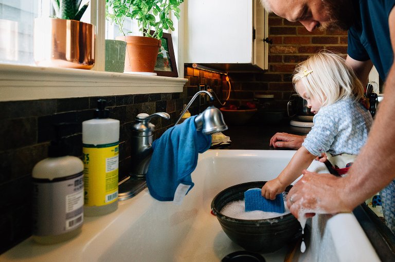 Toddler girl and dad wash a bowl in kitchen sink. 
