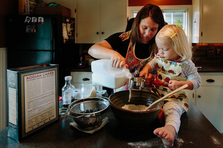 Mom pours milk into measuring cup to make pancakes with toddler daughter. 