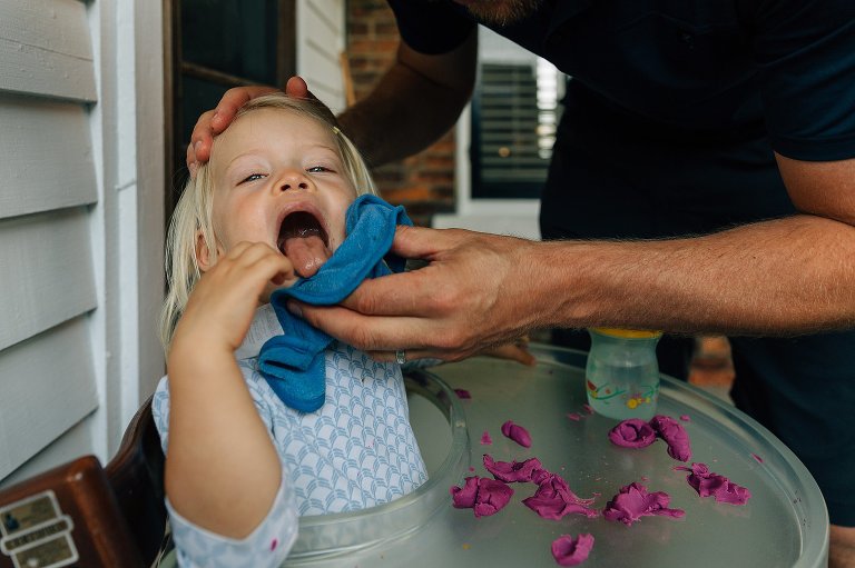 Dad wipes toddler's face of purple Play-dough. Daughter sticks out tongue.