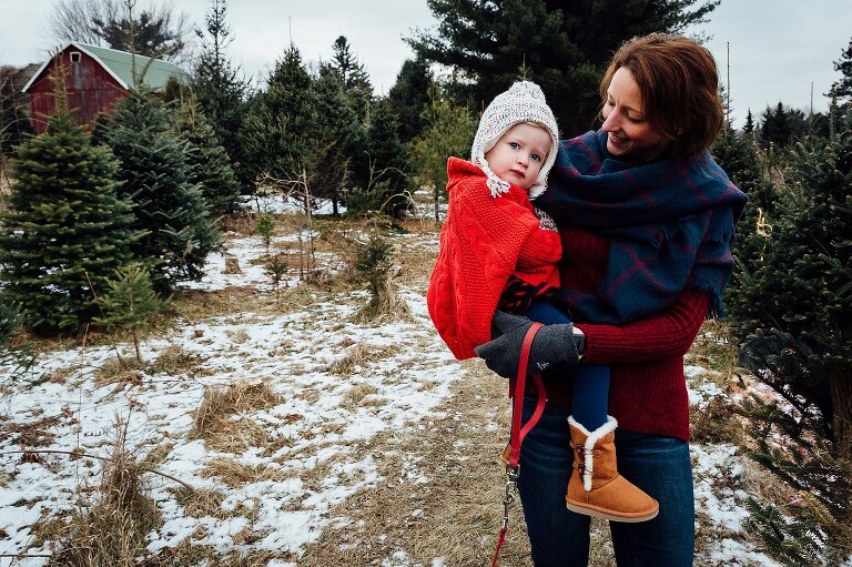a girl in red looks at camera while mother holds her in a field of evergreen trees
