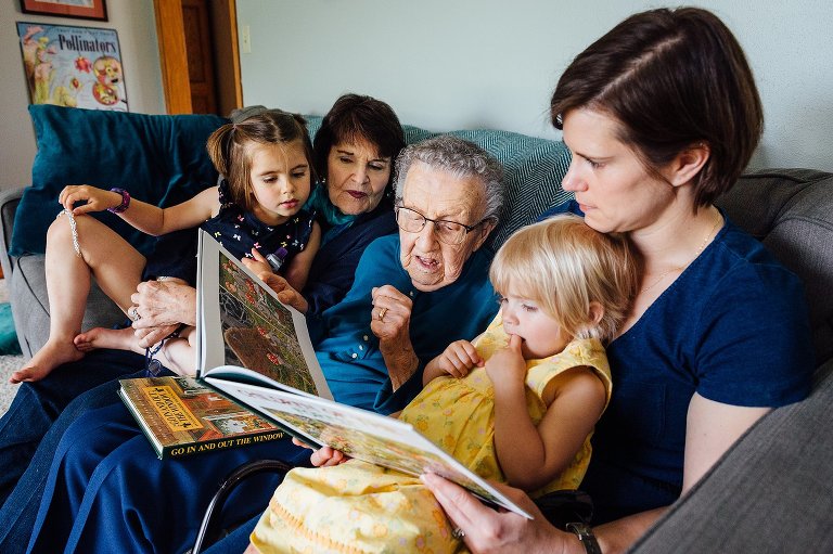 four generations of mothers sit on a couch and read a book to grandchildren