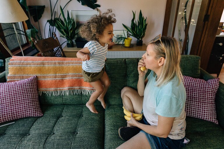 mother holds a banana phone on a couch while young child jumps gleefully on couch