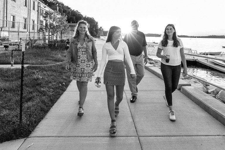 mother walks with two teen girls and spouse near a lake with sunflare present