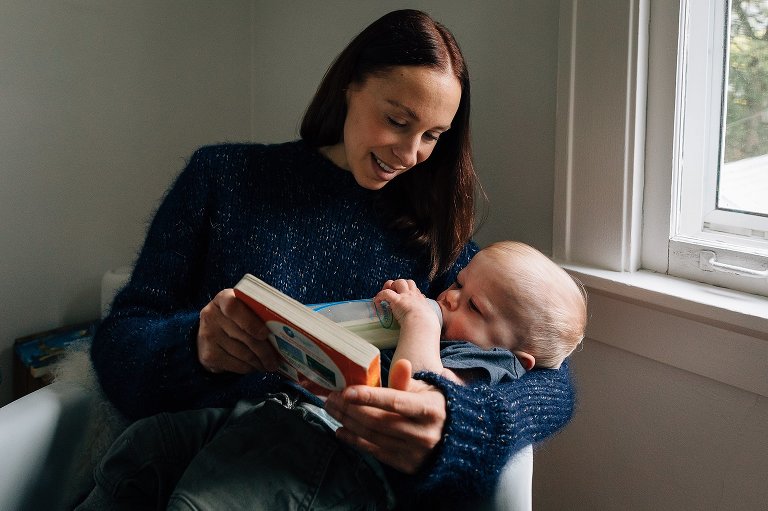 mother holds baby drinking a bottle in a rocking chair while reading a board book