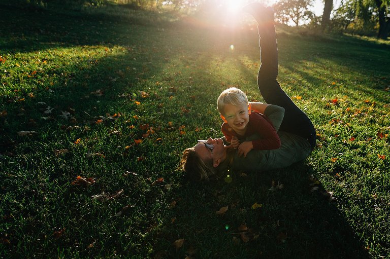 Boy lands on mom after runnning ot her. Smiling. Bright sun flooding frame at top.