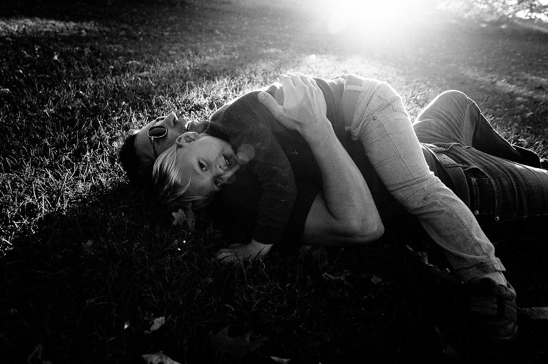 black and white. boy lies on dad in the grass, sunshine flooding frame at top 