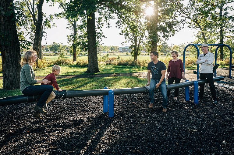 parents, grandparents and small child are on long teeter totter at park. Sun shining through trees. 