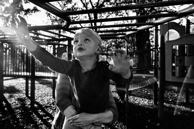 Young boy reaches for next bar on monkey bar playground, mom holds onto his waist 