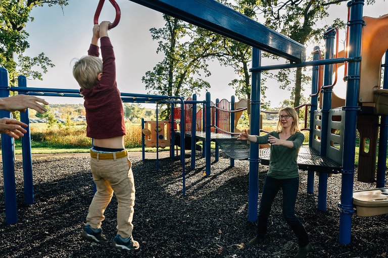 Preschool boy hangs on playground equipment while mom holds out her hands for him. 