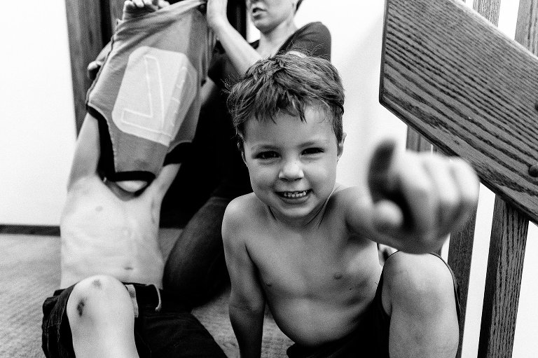 Black and white. Boy points finger at camera as mother helps older brother change clothes before bed. 