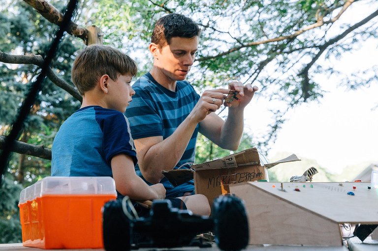 Dad inspects electrical equipment with young son as they work on an invention in a treehouse. 
