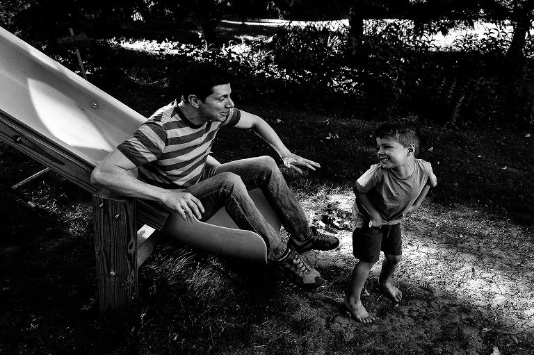 Black and white. Dad slides down slide after young son. They're laughing. 