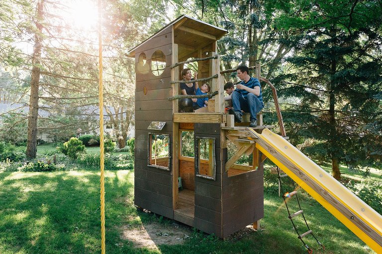 Family sits in a tree house in backyard. Summer evening sun is shining through tall trees. 