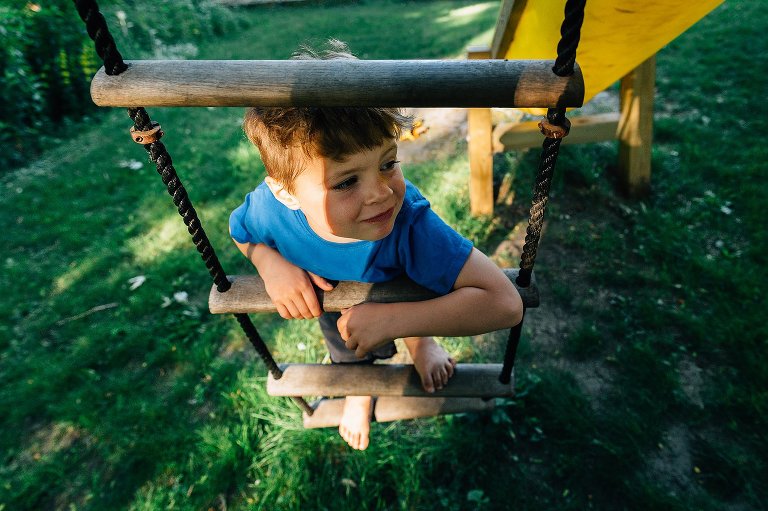 Boy climbs up a rope ladder to his backyard treehouse in the summer. He is barefoot and not camera aware.